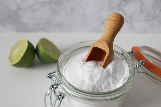 Baking soda, one of the ways how to get rid of german roaches, in a glass bowl with a brown, wooden scoop. In the background of the image sits green limes. 