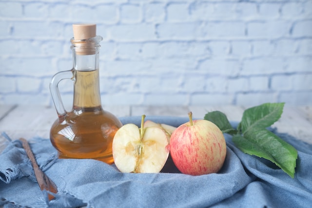 Bottle of apple cider vinegar beside two apples in a blue basket against a white brick wall 