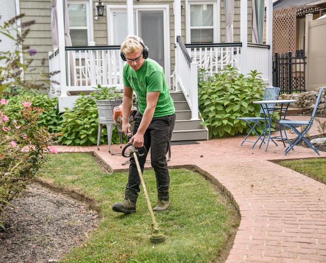 Blond man with green shirt and black pants doing yard work in front of a white and grey house. The man is weed eating with headphones on and there is evidence of other yardwork that has been completed, a way how to deter stink bugs